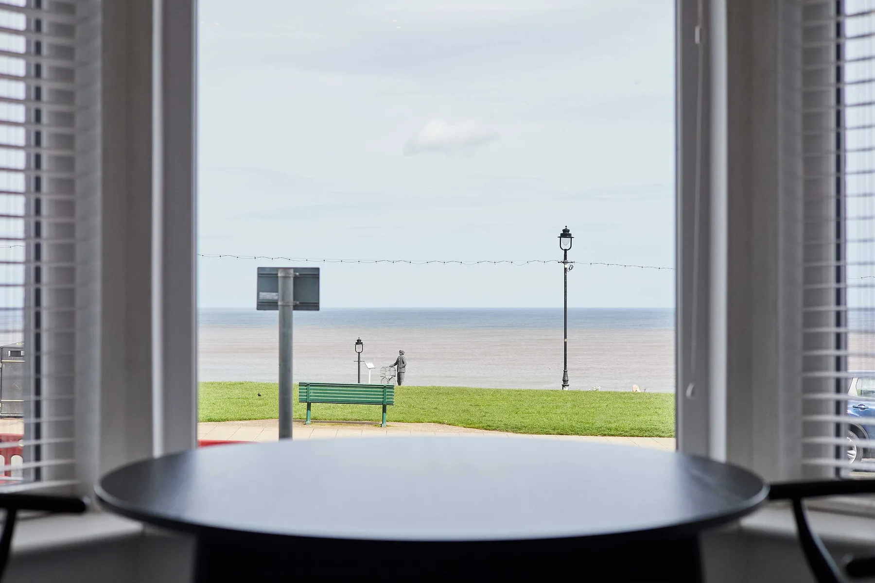 A round table faces a seaside promenade through an open window.
