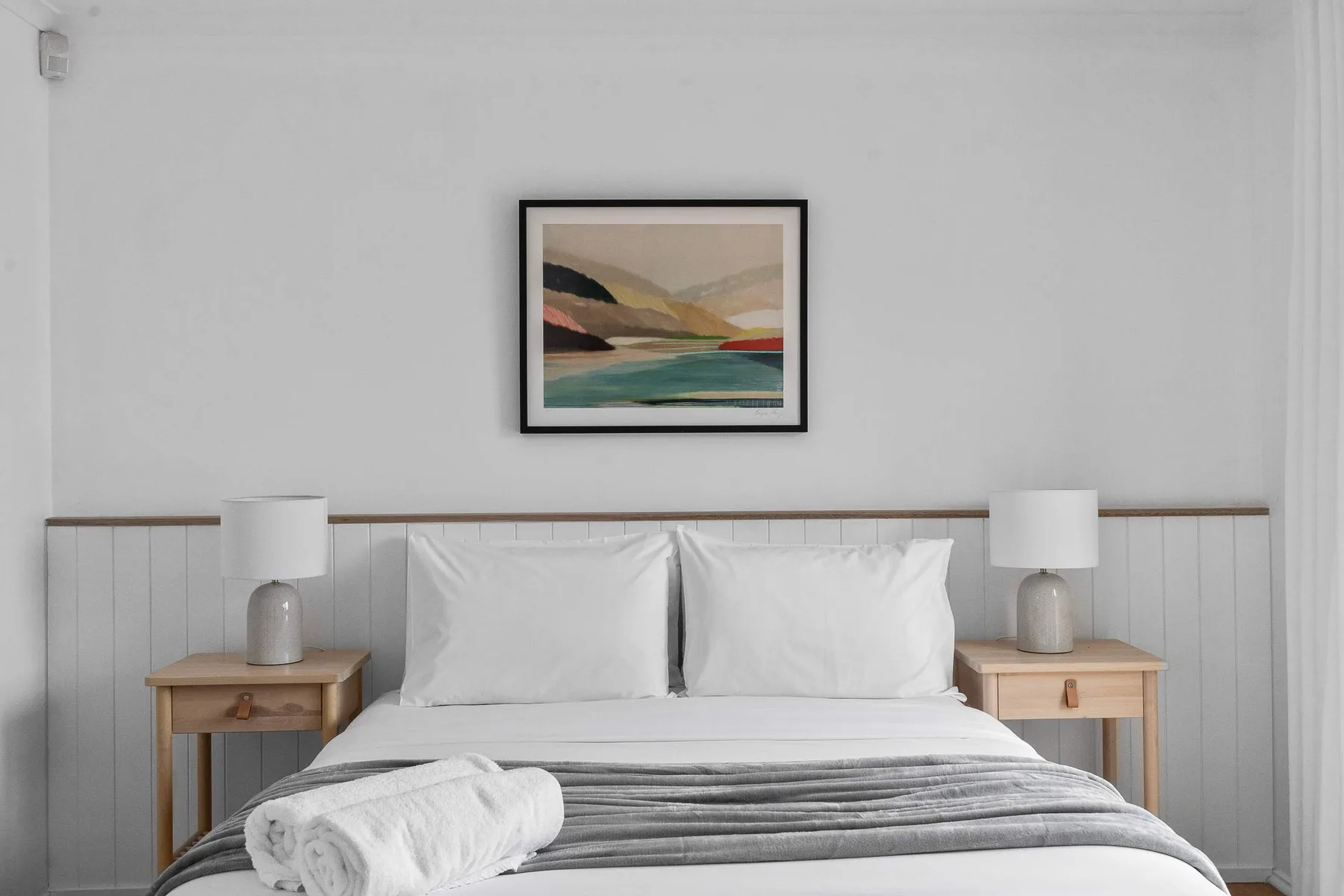 A bedroom with a neatly made bed, bedside tables, and a framed landscape print.
