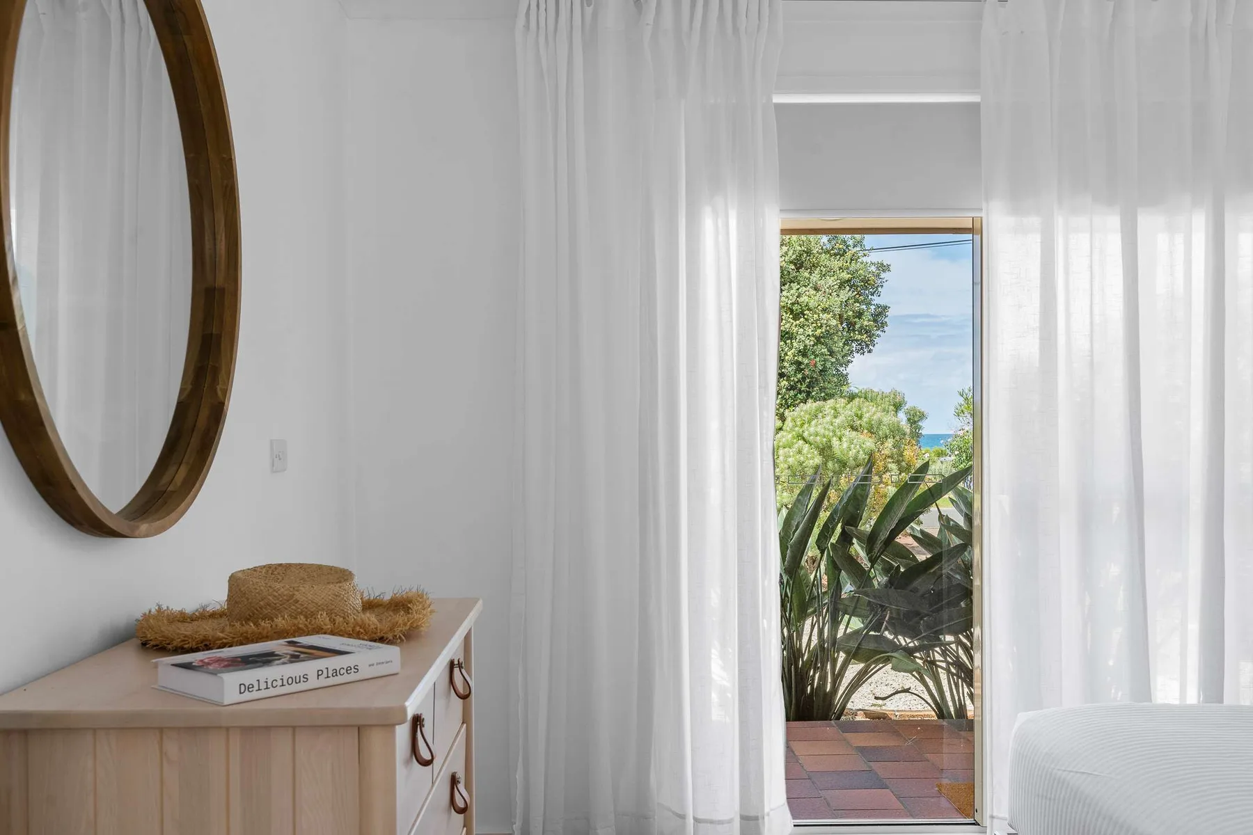 A bright bedroom with sheer curtains and a view of leafy plants through the glass door.
