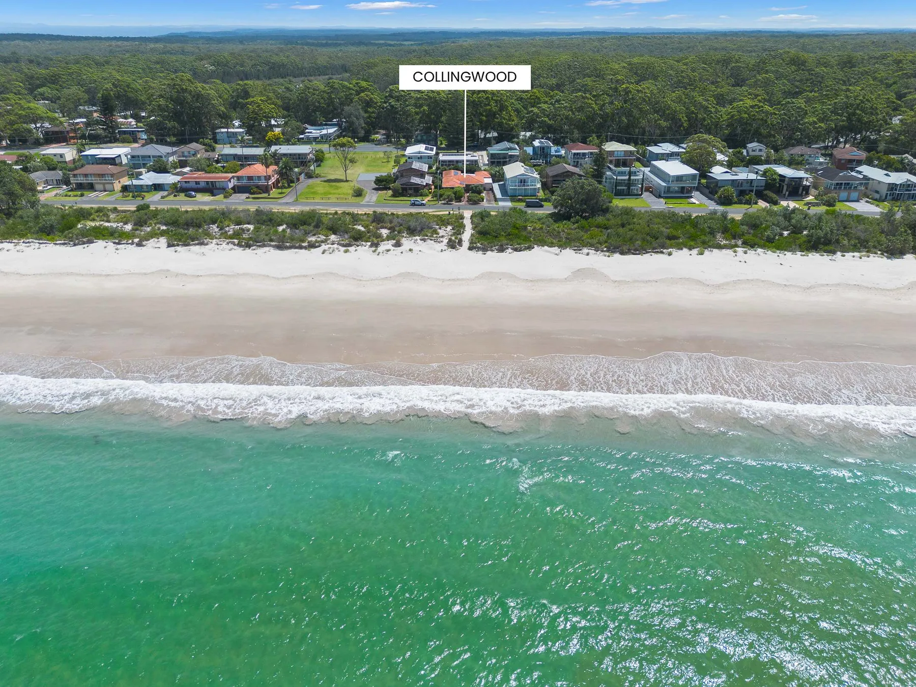 An aerial view of a sandy beach, coastal homes, and green ocean water.
