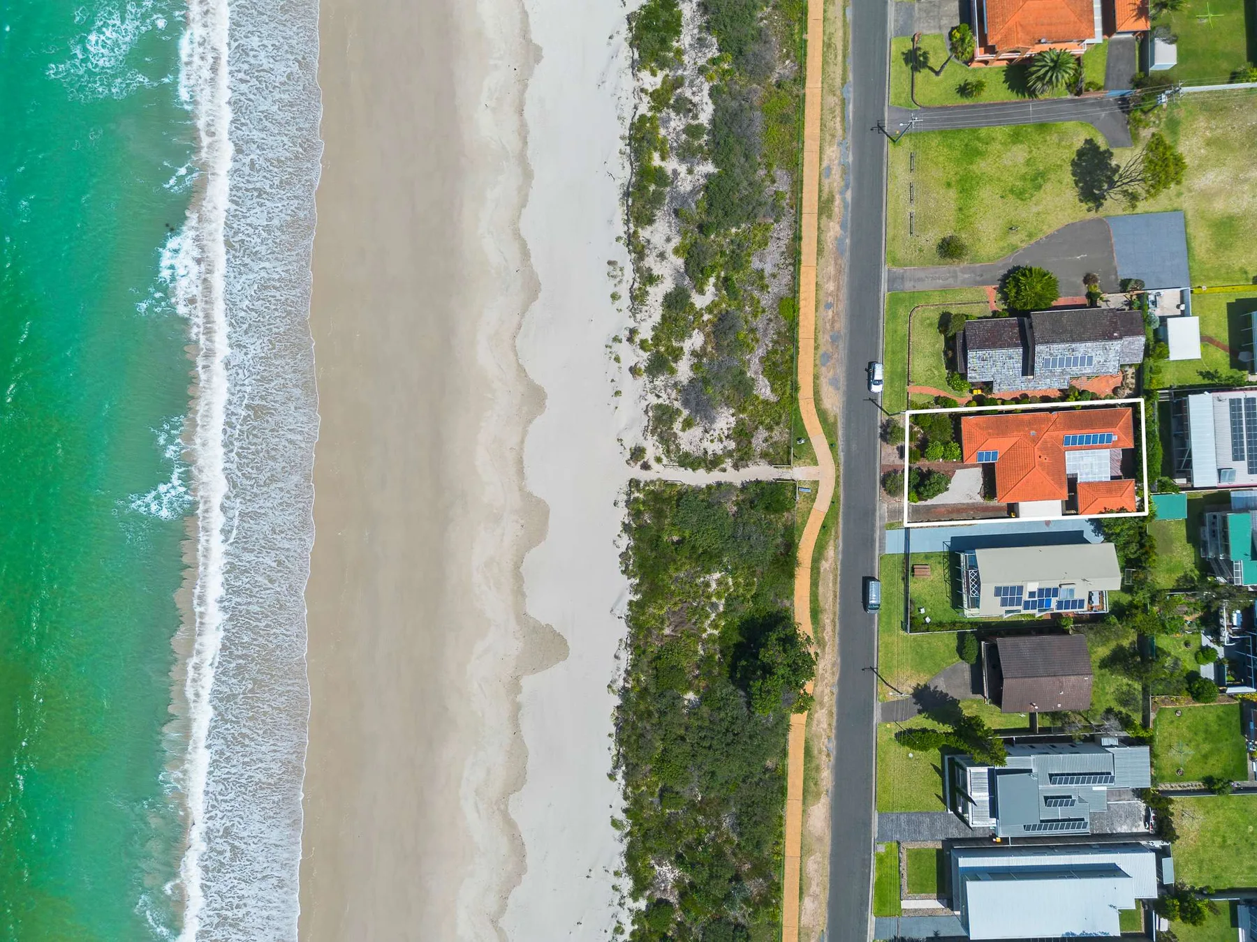 An aerial view shows a sandy beach beside coastal homes and a road.