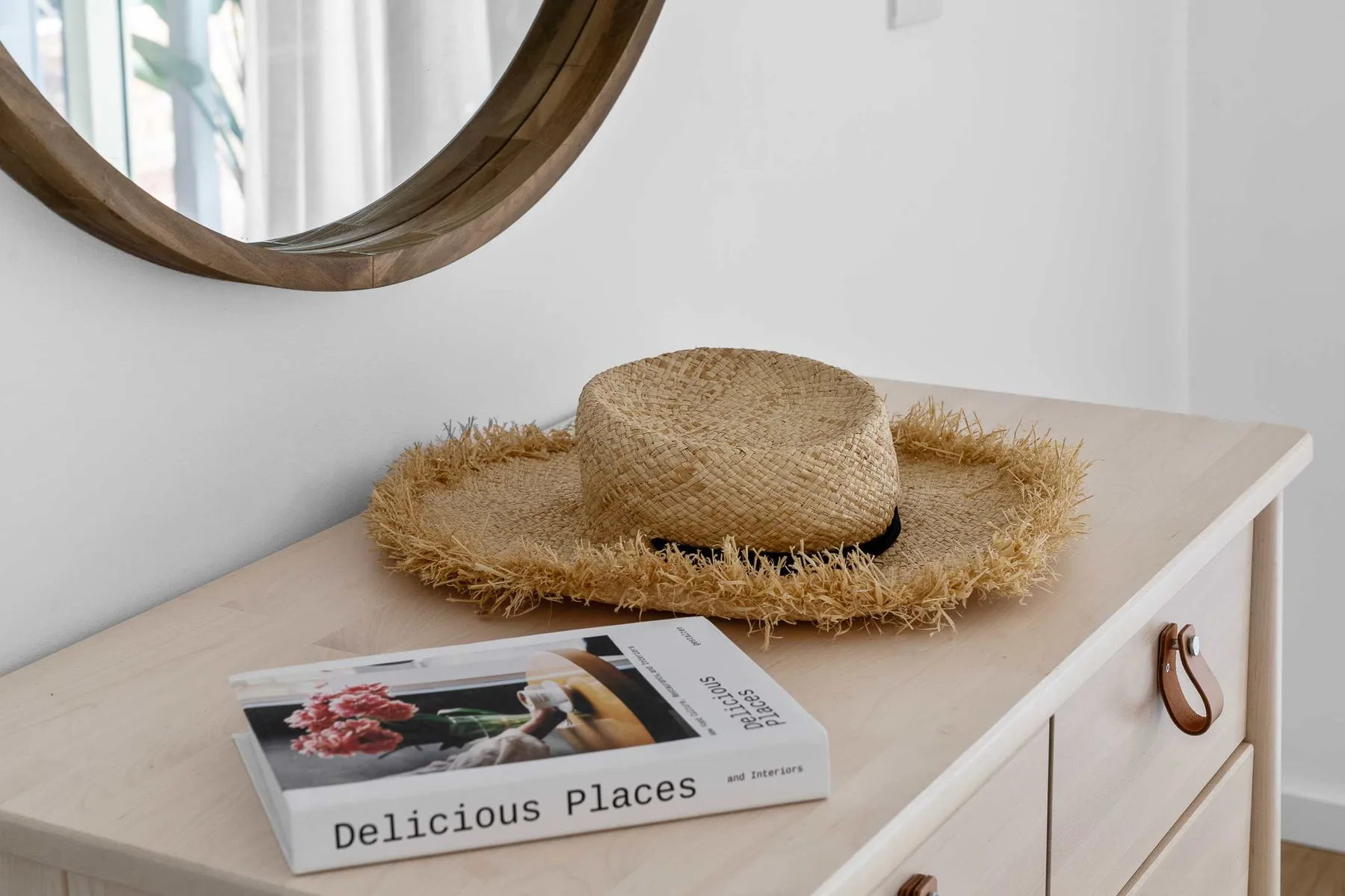 A light-colored dresser holds a straw hat and a book beneath a round mirror.