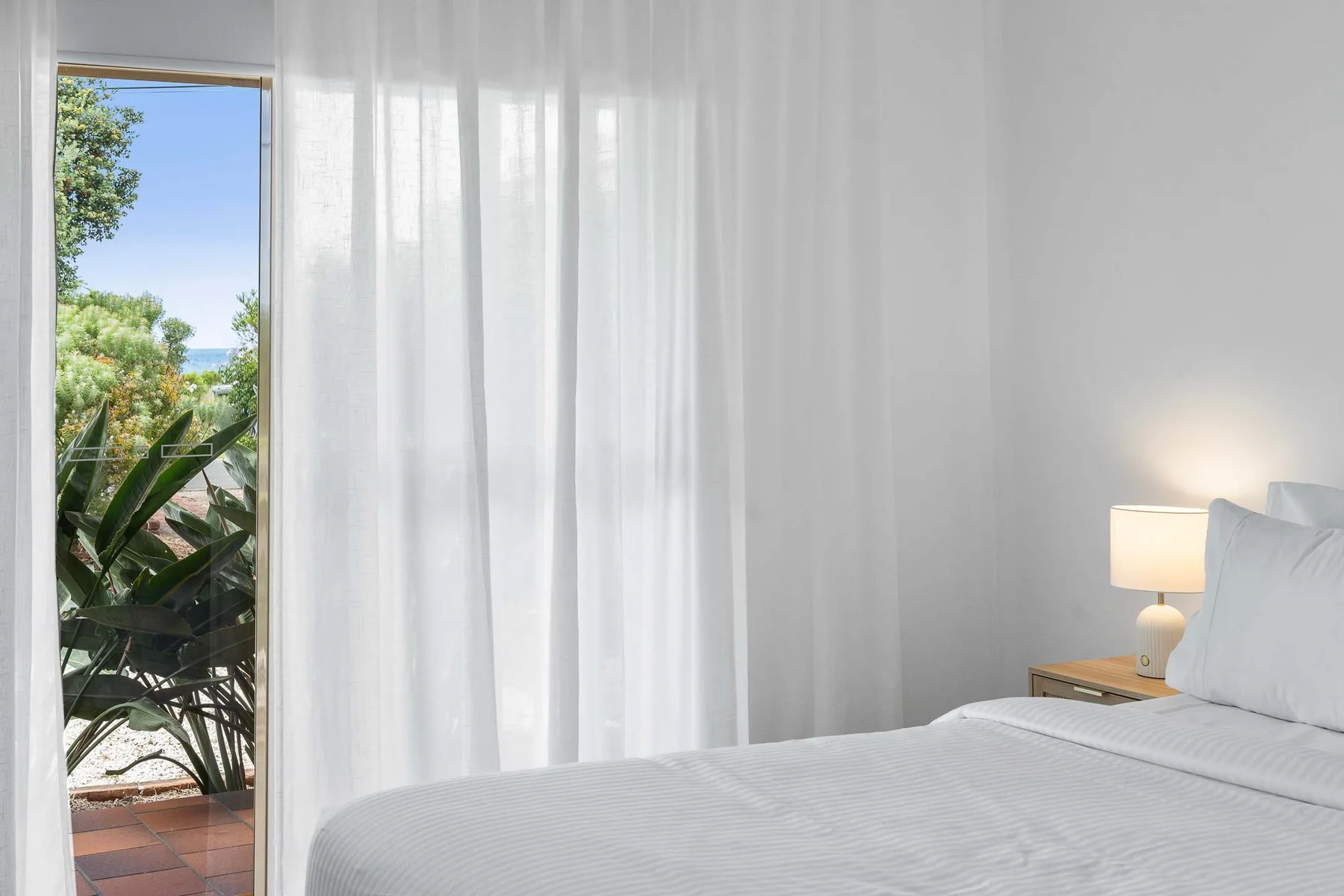 A bright bedroom with a bed beside sheer curtains overlooking greenery.