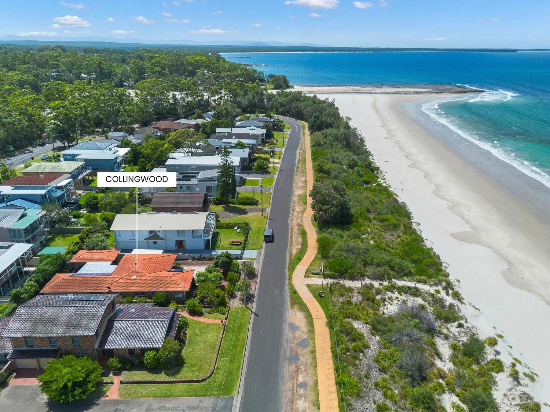 Aerial view of coastal homes beside a road, sandy beach, and blue water.