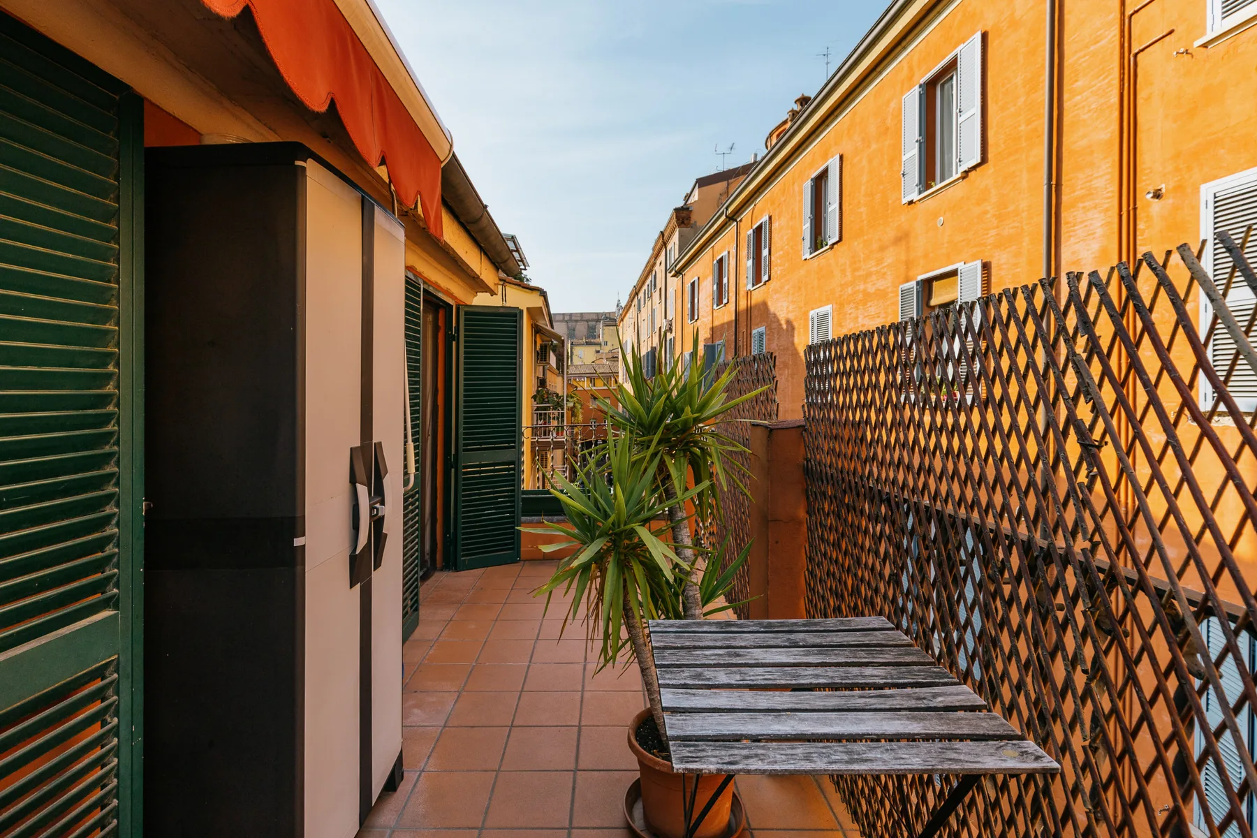 Una terraza exterior estrecha con una mesa pequeña, plantas en macetas y una celosía.