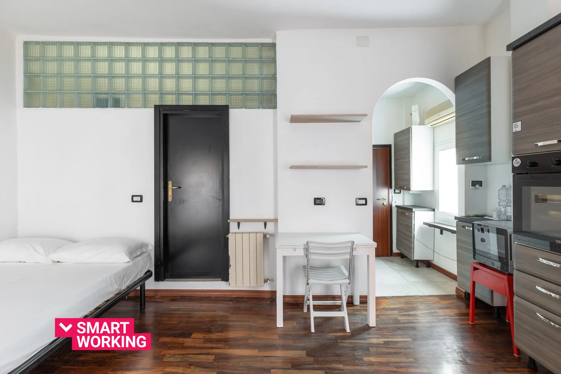 Una habitación tipo estudio luminosa tiene una cama, una mesa pequeña y una zona de cocina.