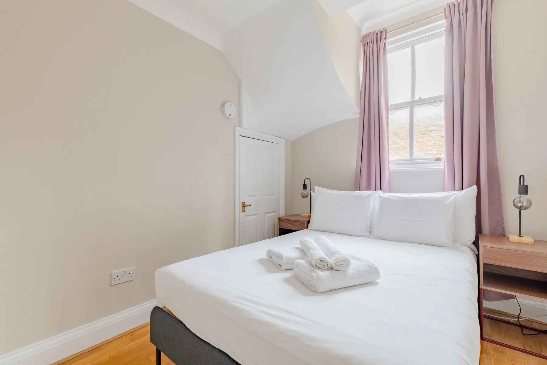 A bright bedroom with a neatly made bed beneath a window with pink curtains.