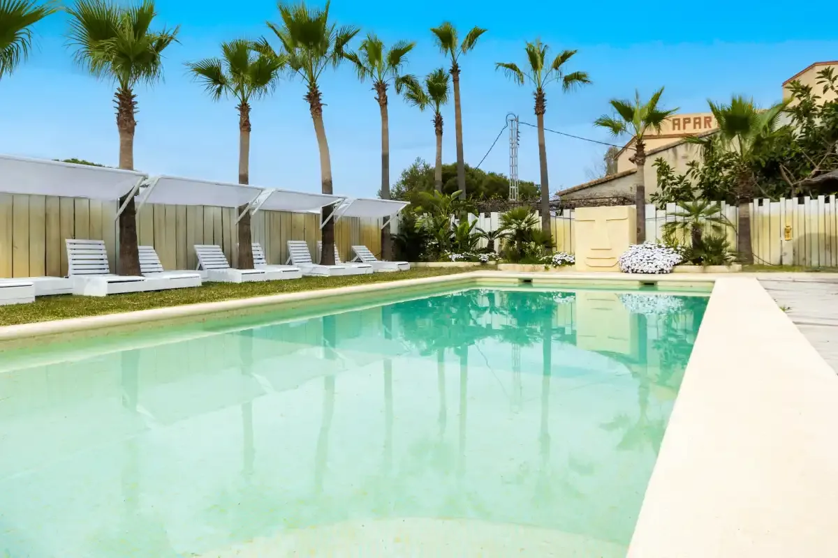 Una piscina rodeada de palmeras y cabañas con sombra.