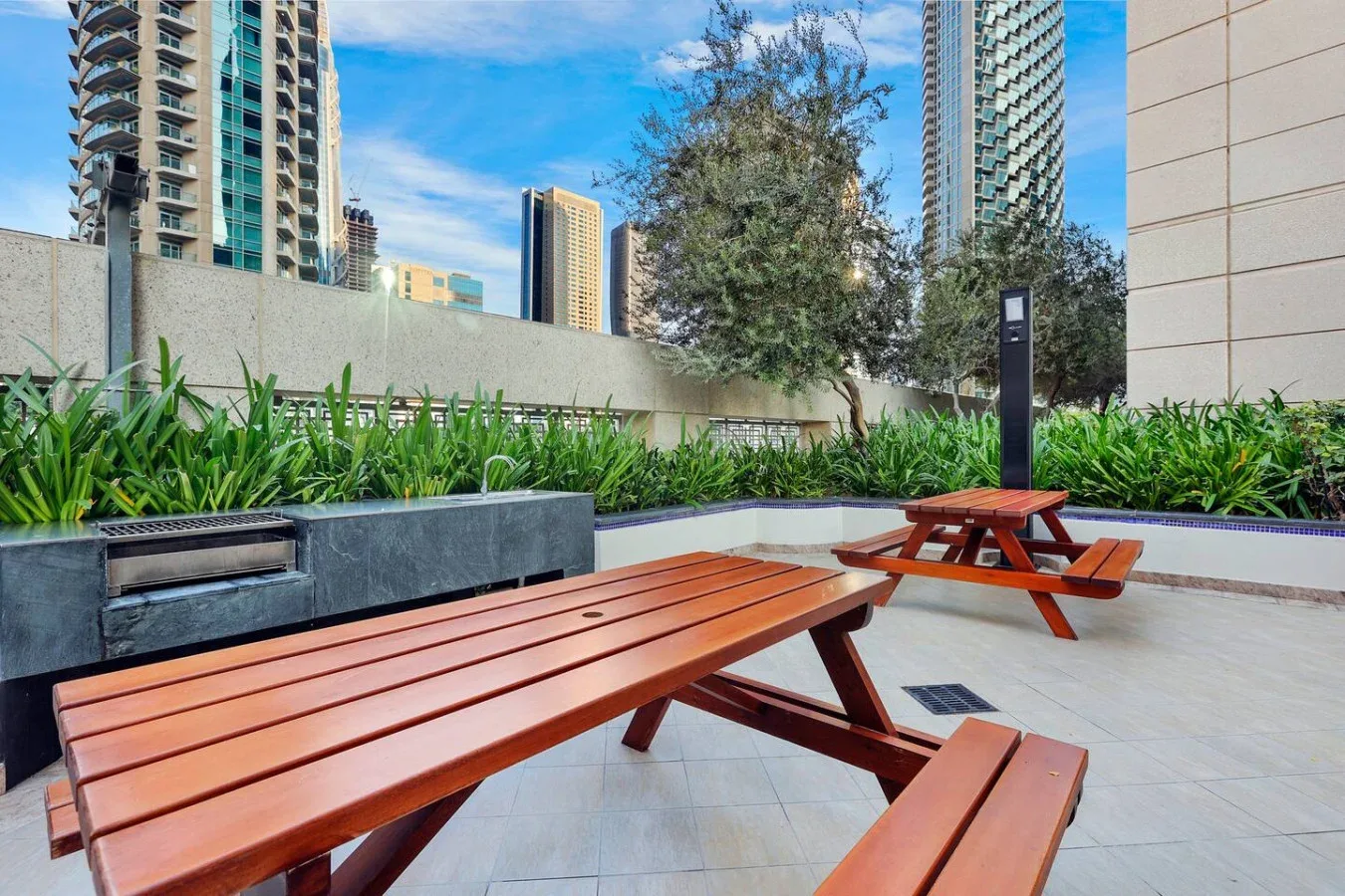 Un patio al aire libre con mesas de picnic de madera, jardineras y edificios altos al fondo.