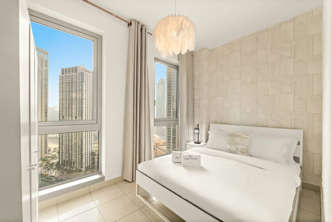 Un dormitorio luminoso con una cama blanca junto a ventanas altas con vistas a edificios de la ciudad.