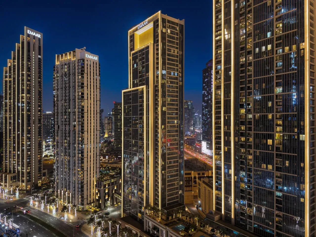 Un paisaje urbano nocturno con edificios altos iluminados junto a una avenida transitada.