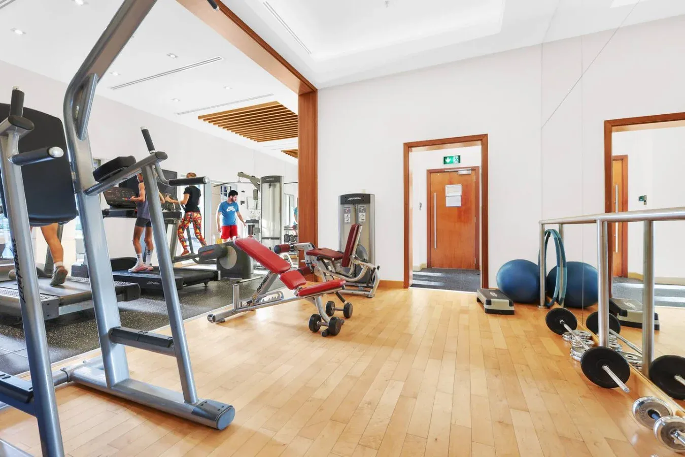 Un gimnasio con máquinas de ejercicio, pesas y suelo de madera clara.