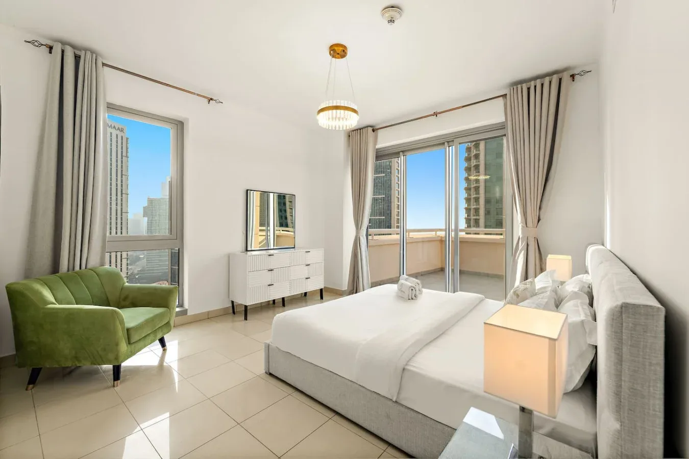 Un dormitorio luminoso con una cama, asientos verdes y grandes ventanas que dan a un balcón.
