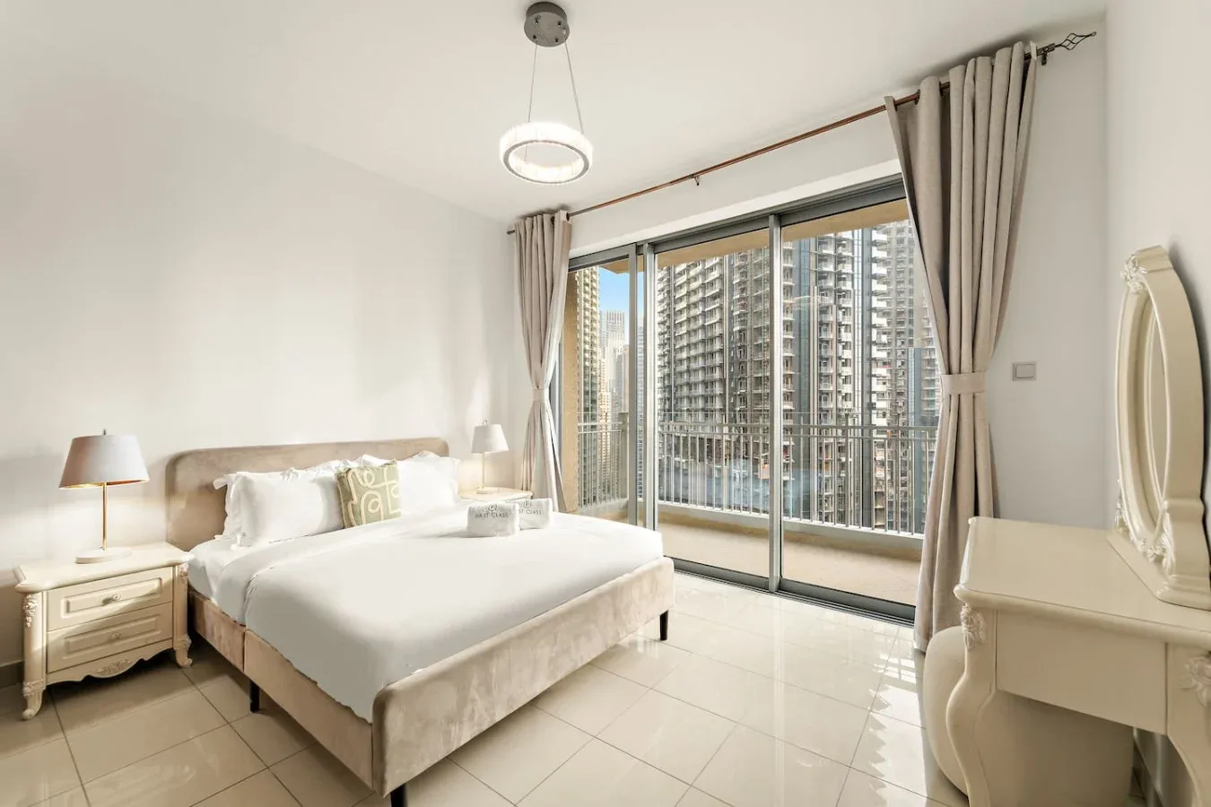 Un dormitorio luminoso con una cama junto a grandes ventanales que dan a edificios altos.