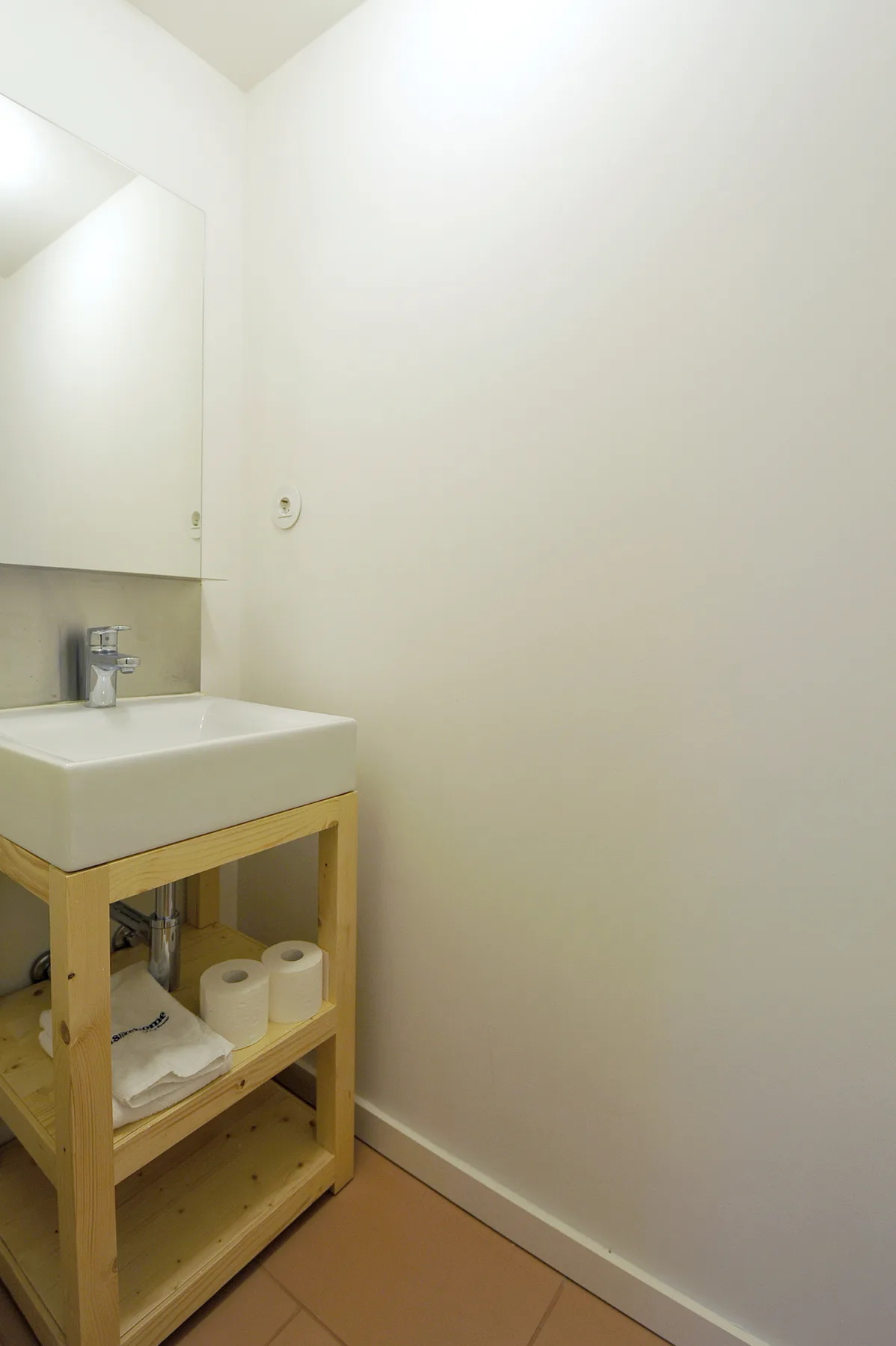 A small bathroom has a sink on a wooden stand beside a plain wall.