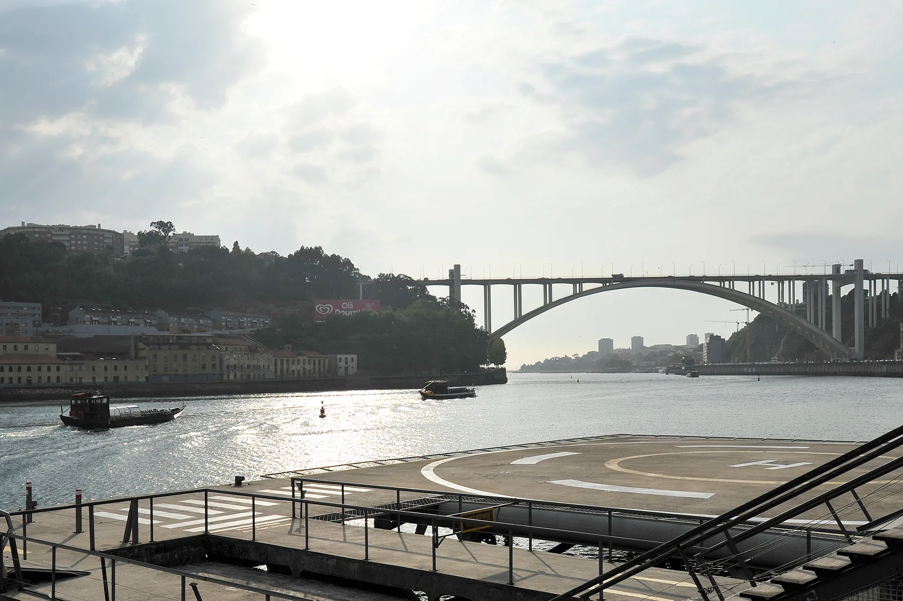 A riverside waterfront with boats on the water and a large arched bridge in the distance.