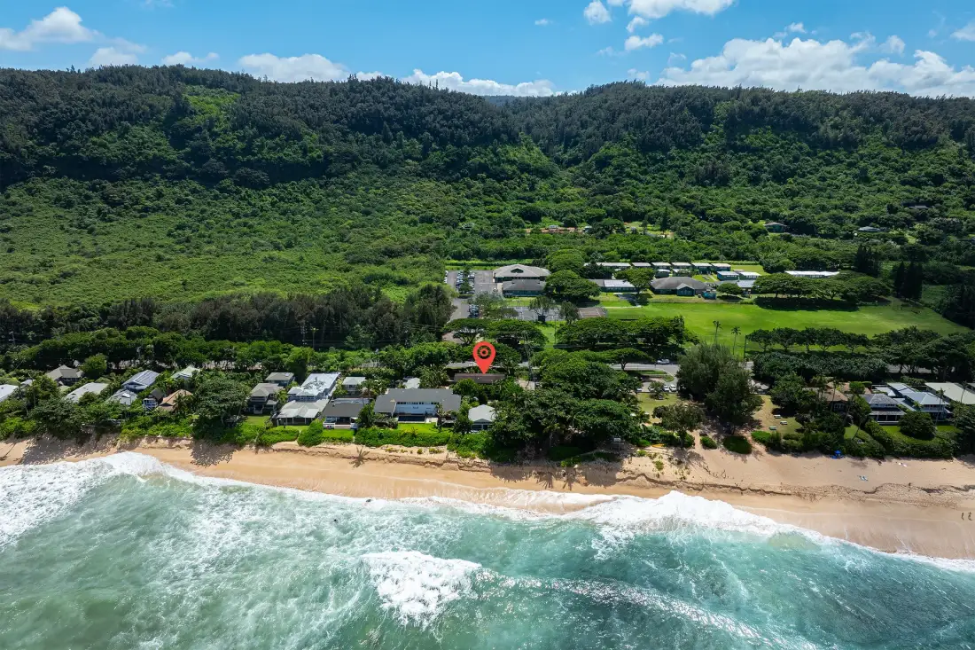 Vista aérea de un asentamiento costero verde junto a una playa de arena y colinas boscosas.