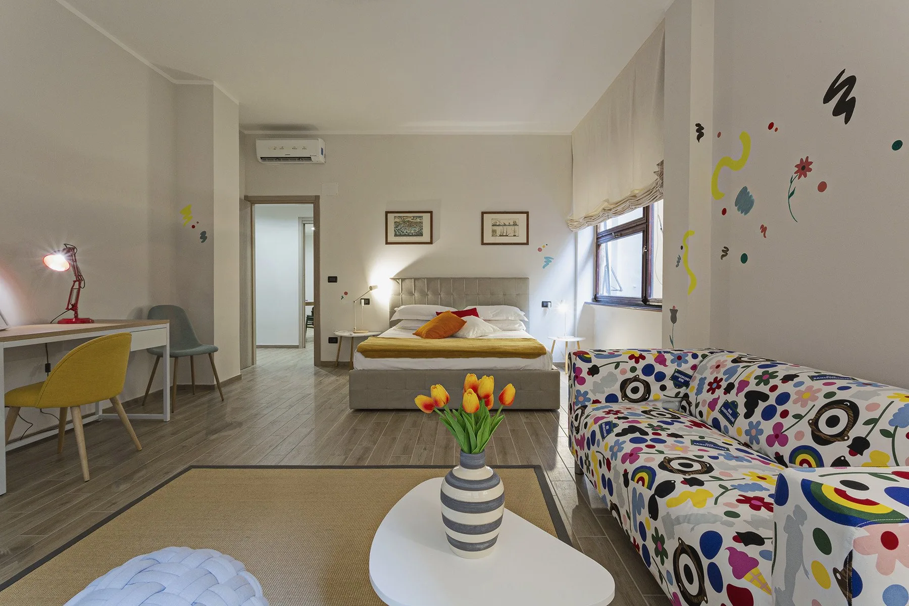 A bright bedroom with a bed, colorful seating, and a small dining area.