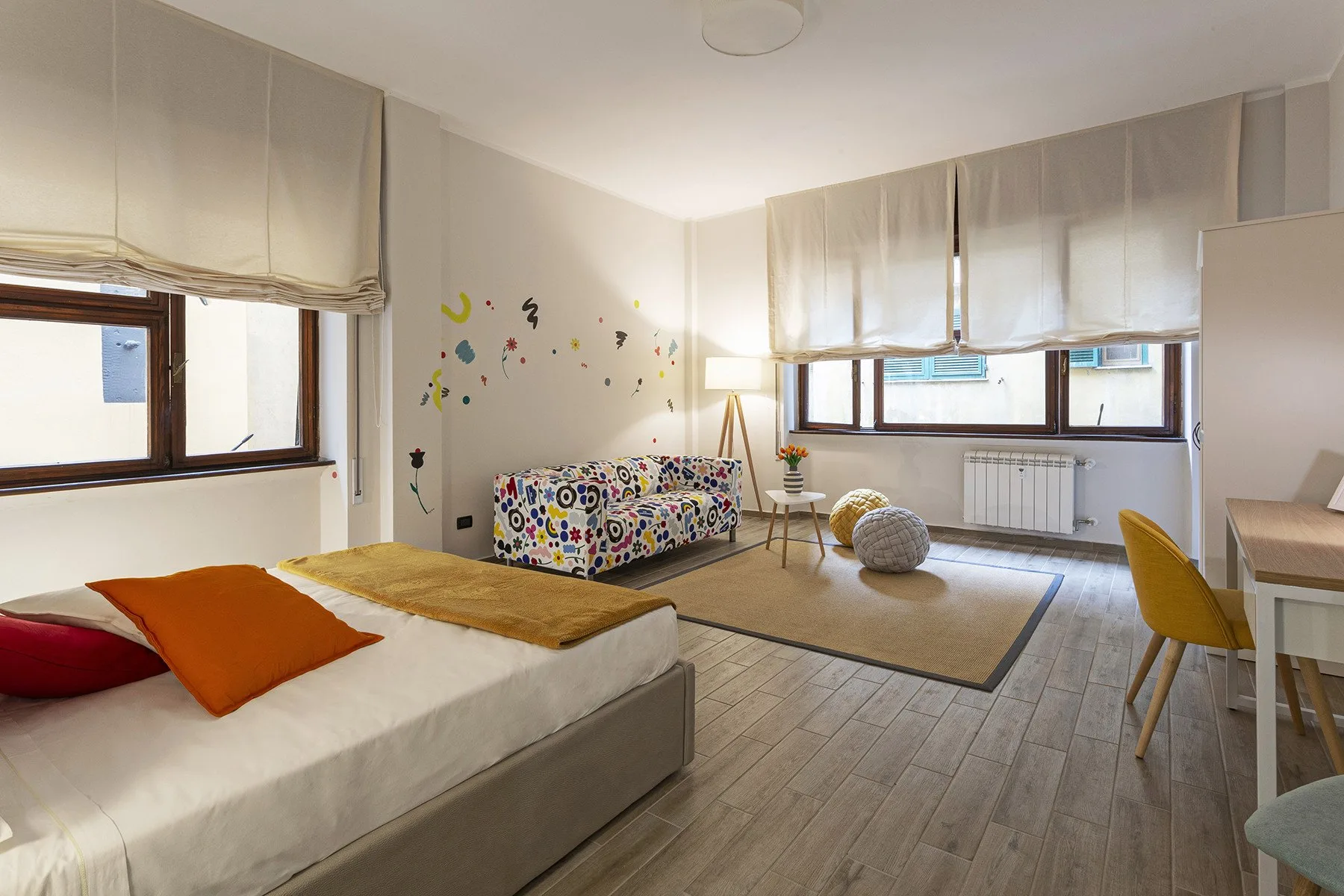 A bright bedroom with a bed, a colorful sofa, and large windows.