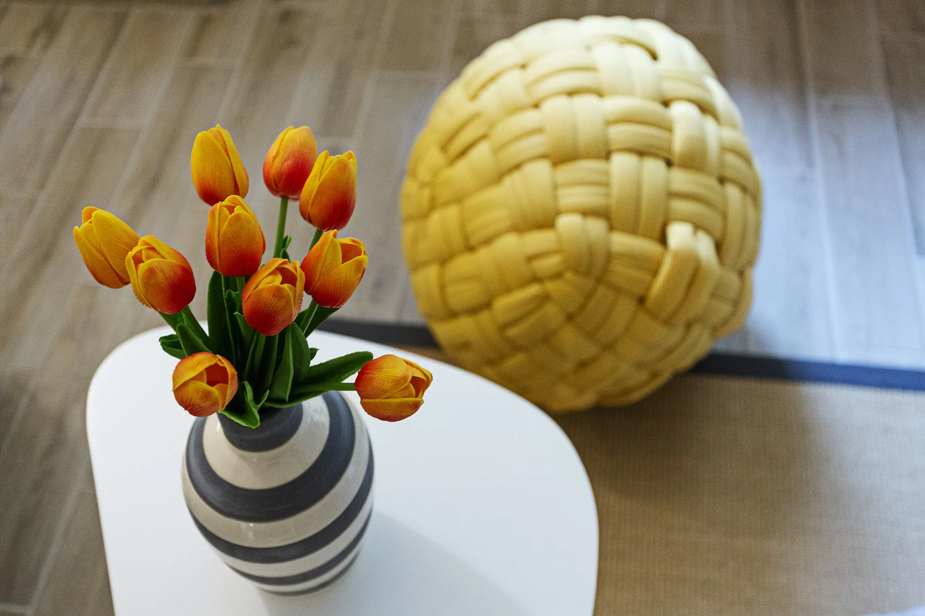 A white table holds a striped vase of orange tulips beside a woven round object.