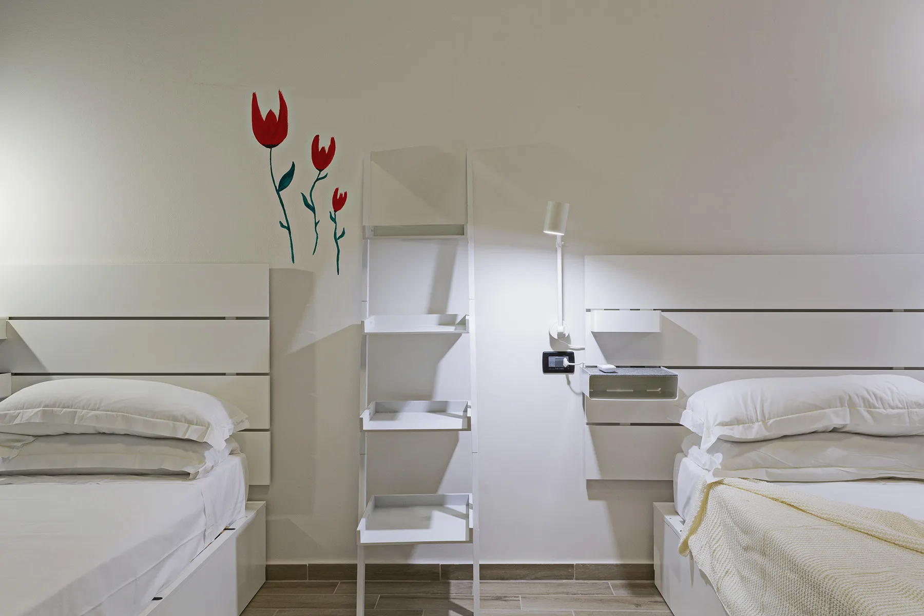 A bright bedroom with two beds and a narrow shelving unit between them.