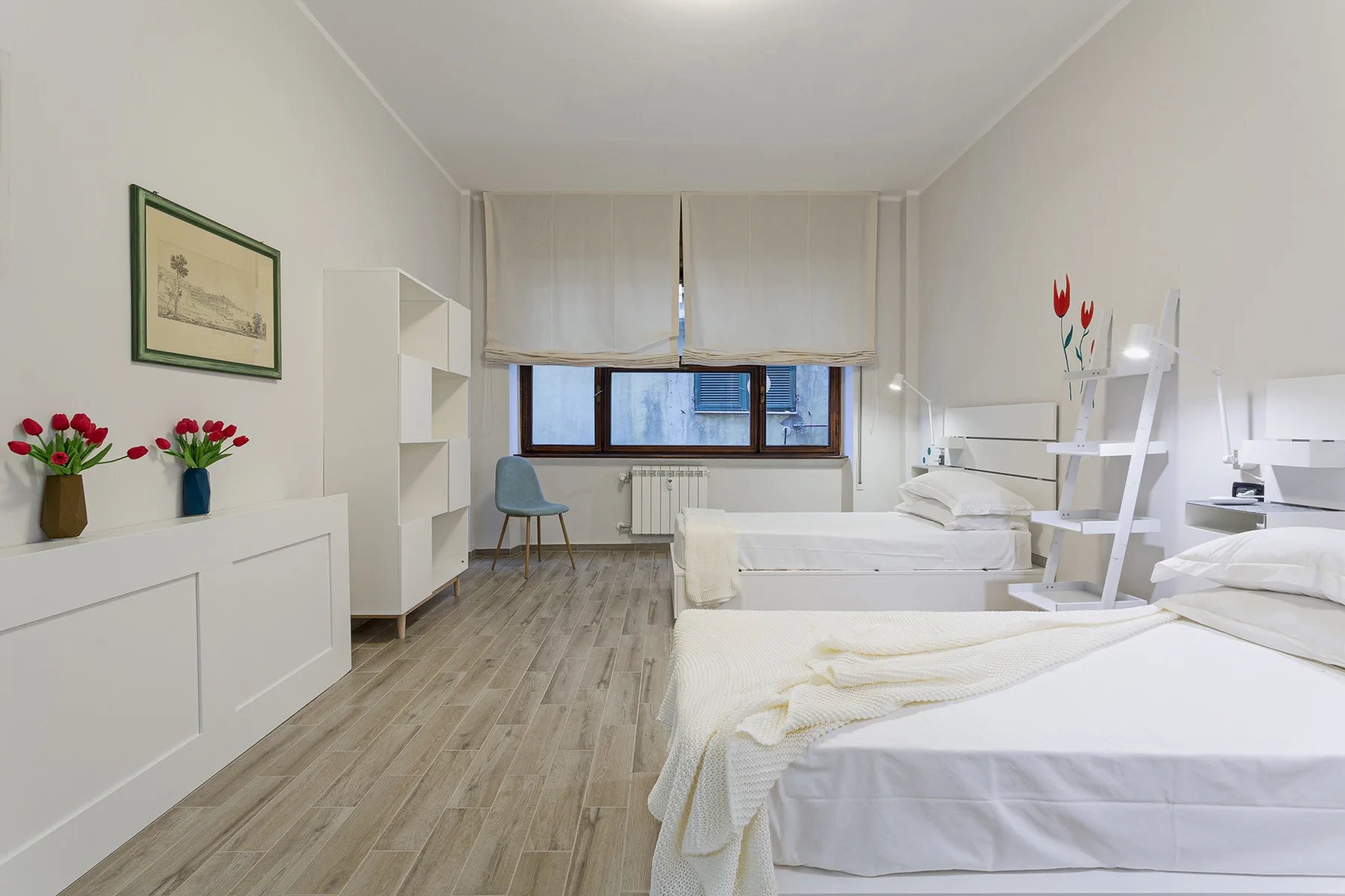 A bright bedroom with two beds, a desk, and a large curtained window.