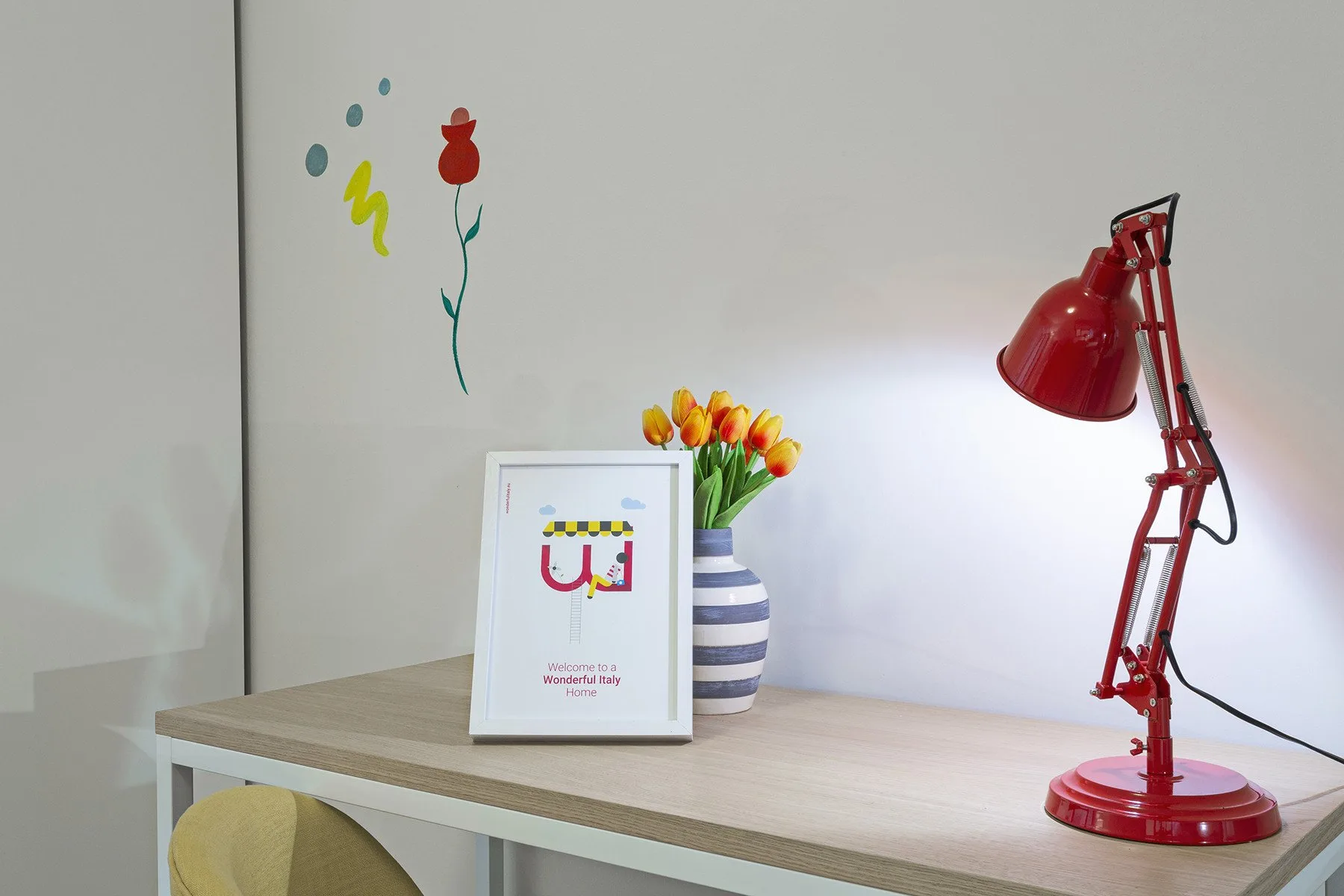 A desk displays a red lamp, flowers, and a framed print against a decorated wall.