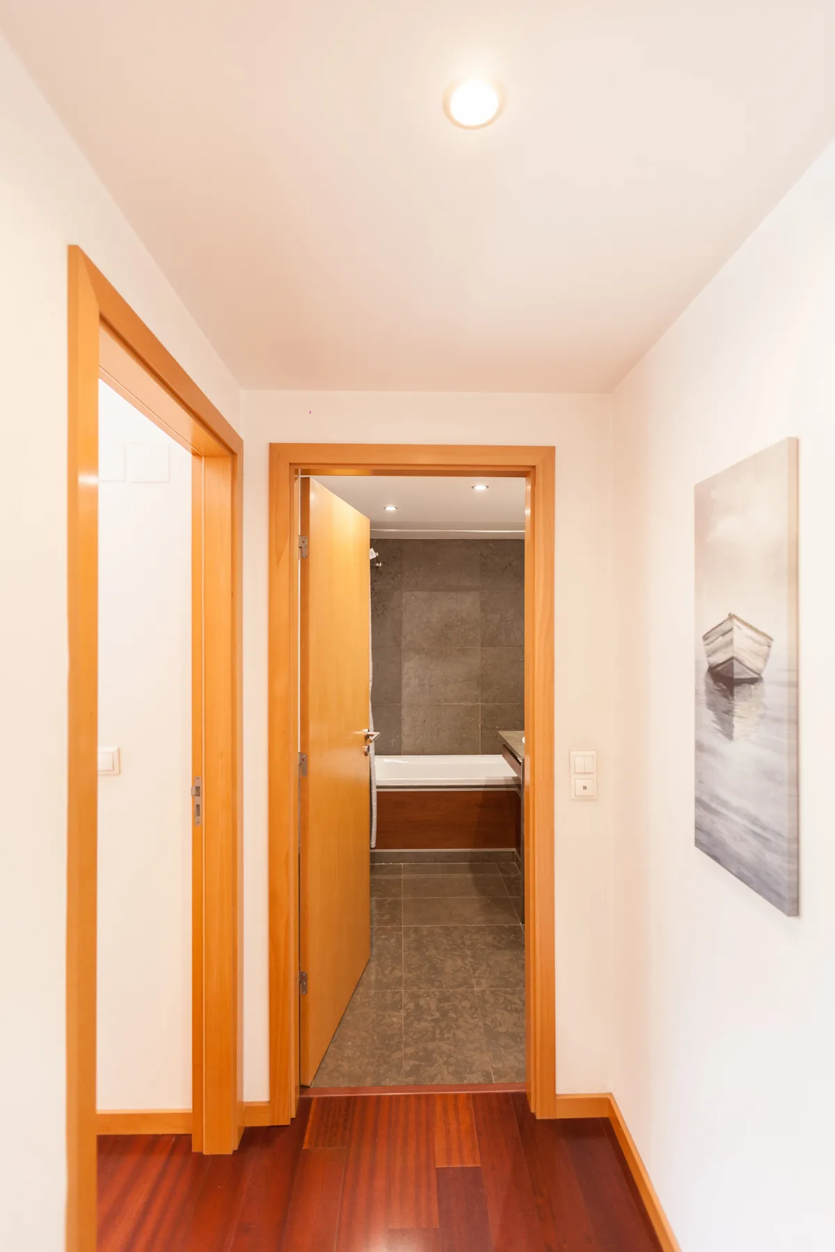 A hallway with wooden flooring leads to a bathroom with a bathtub.