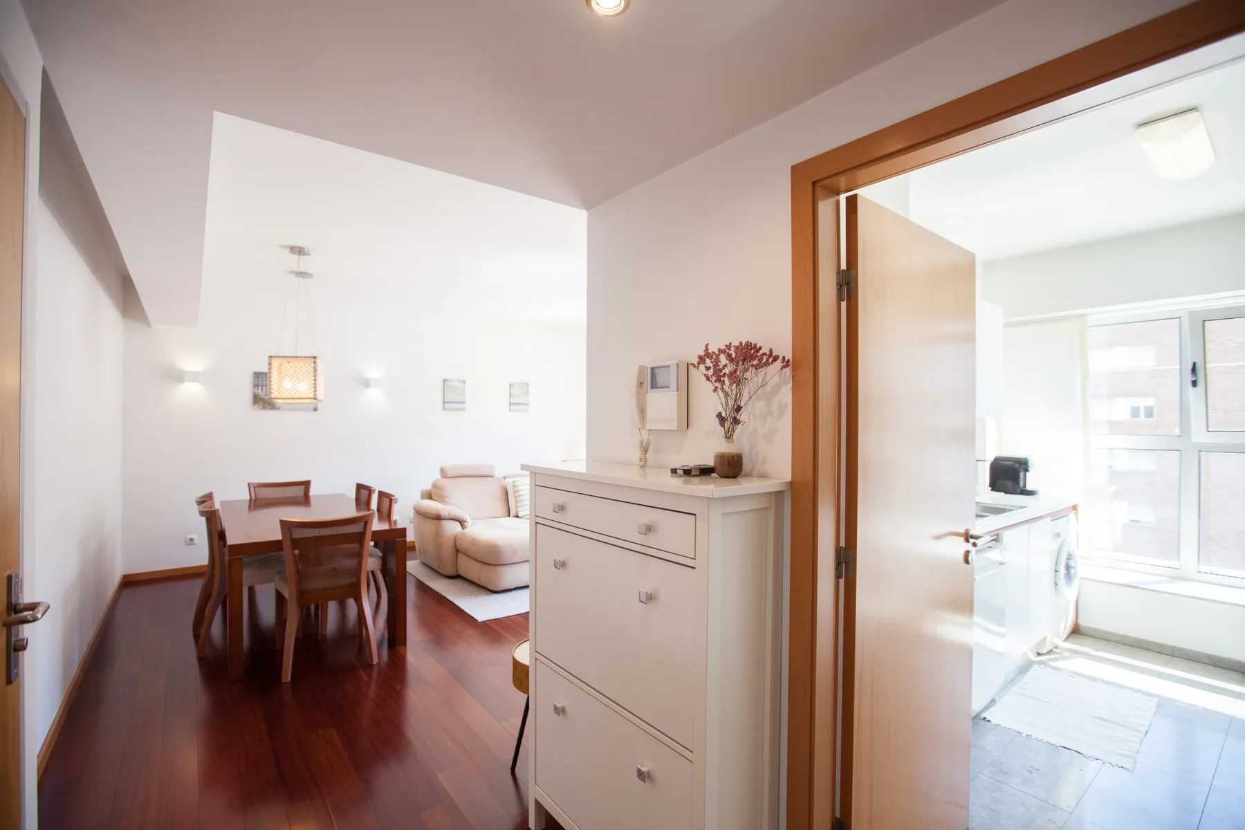 A bright apartment interior with a dining area, seating, and an open doorway to a sunlit room.