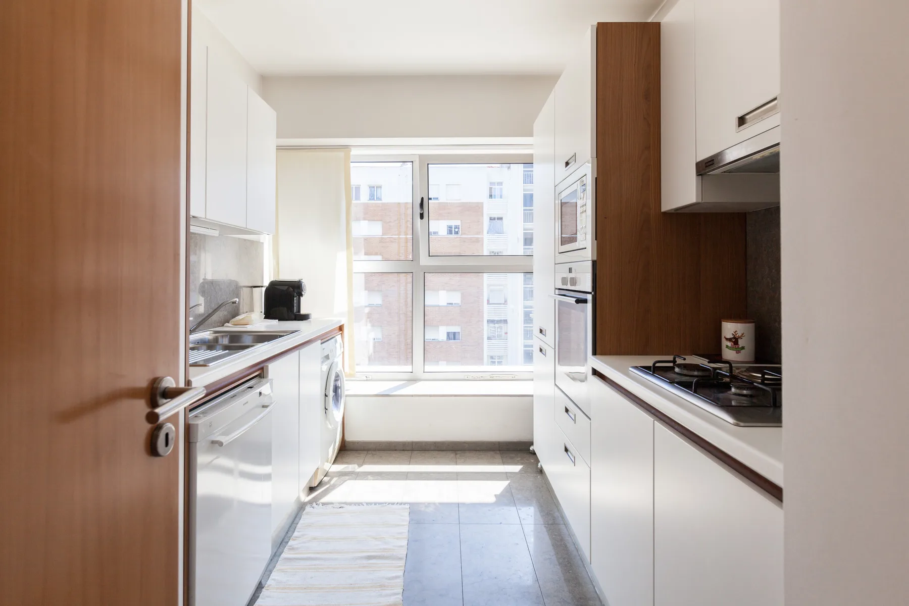 A narrow kitchen with white cabinets and a large window at the far end.