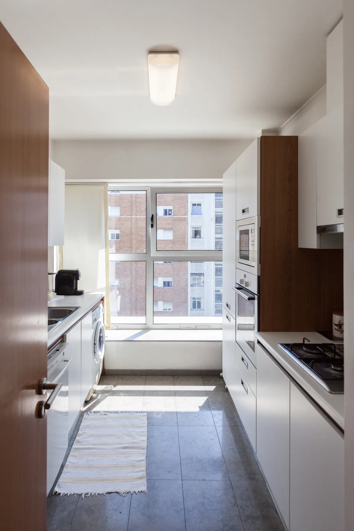 A bright galley kitchen with white cabinets, appliances, and a large window.