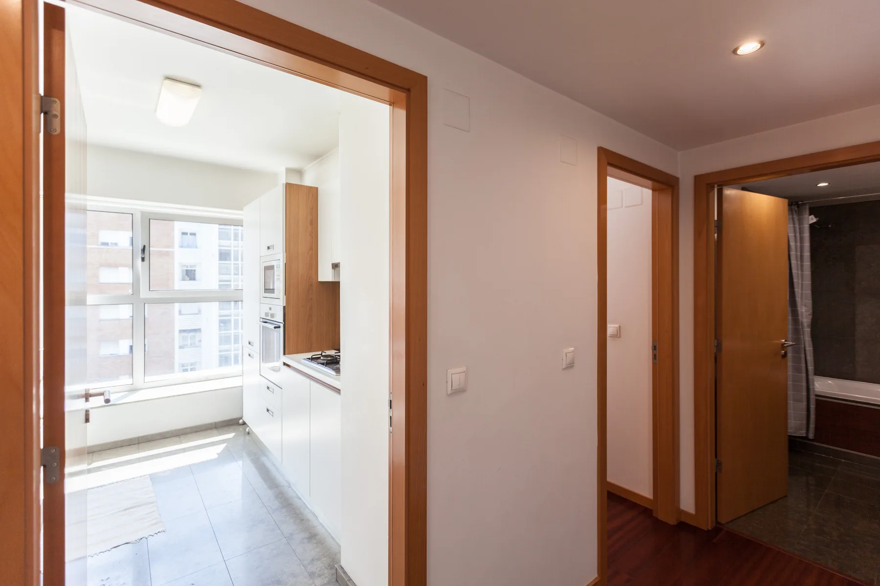 A hallway with open doorways leads to a bright kitchen with large windows.