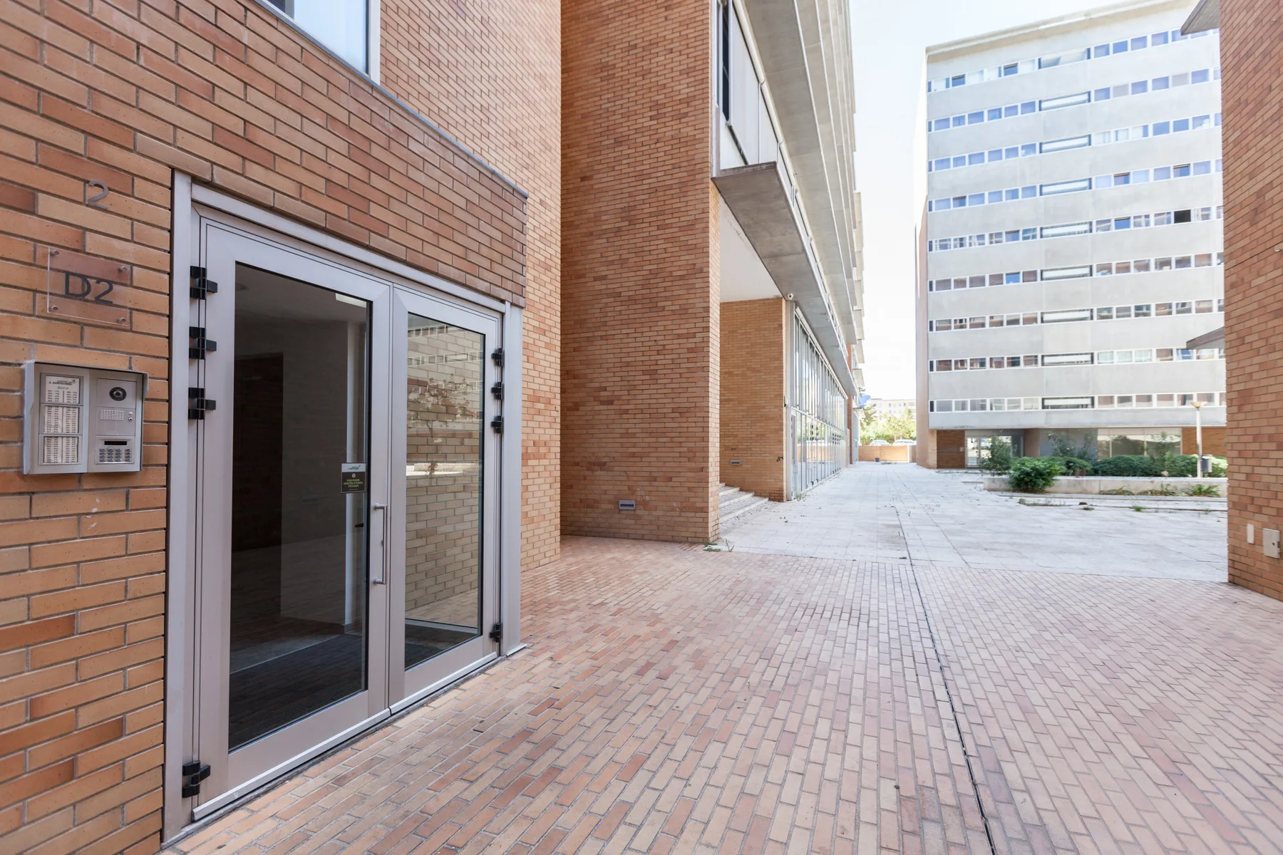A brick-paved courtyard runs between brick buildings, with a glass entrance door.