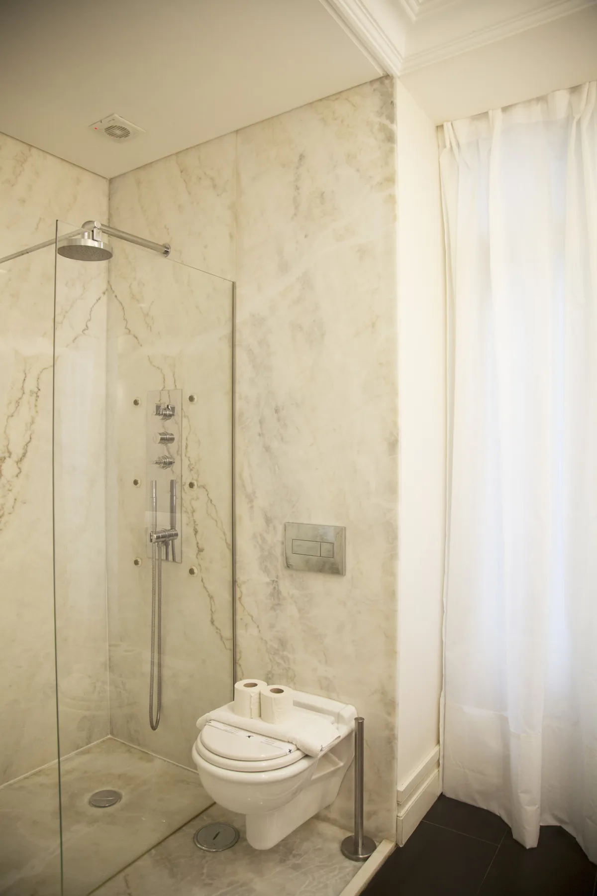 A bathroom with a glass shower and a toilet beside a curtained window.