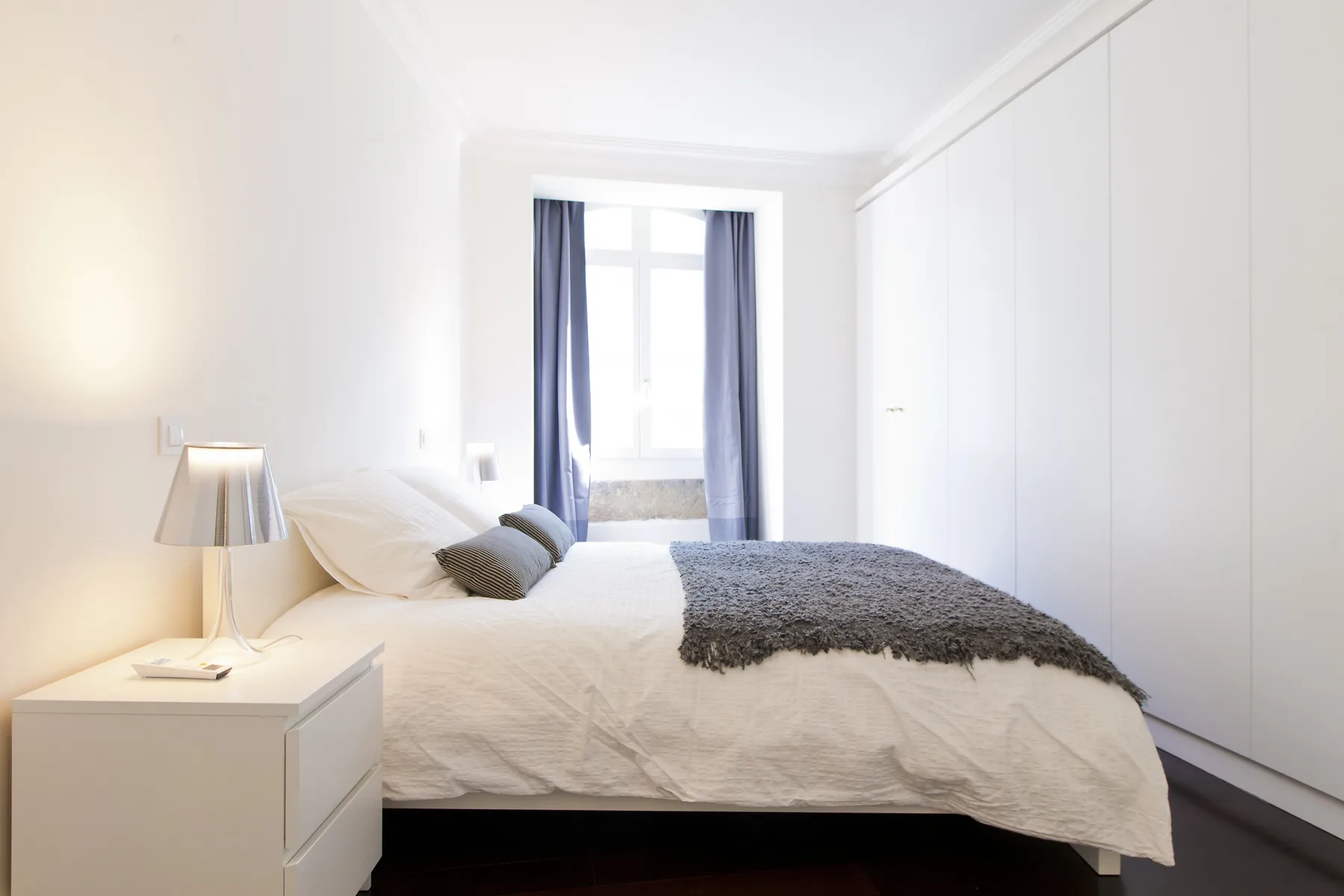 A bright bedroom with a bed, bedside table, and window with curtains.