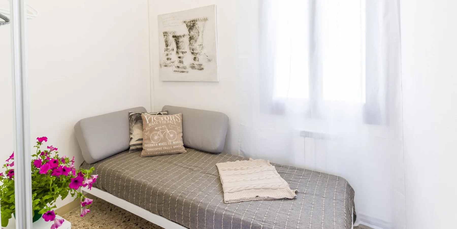Una cama de día está bajo una lámina enmarcada junto a una ventana luminosa.