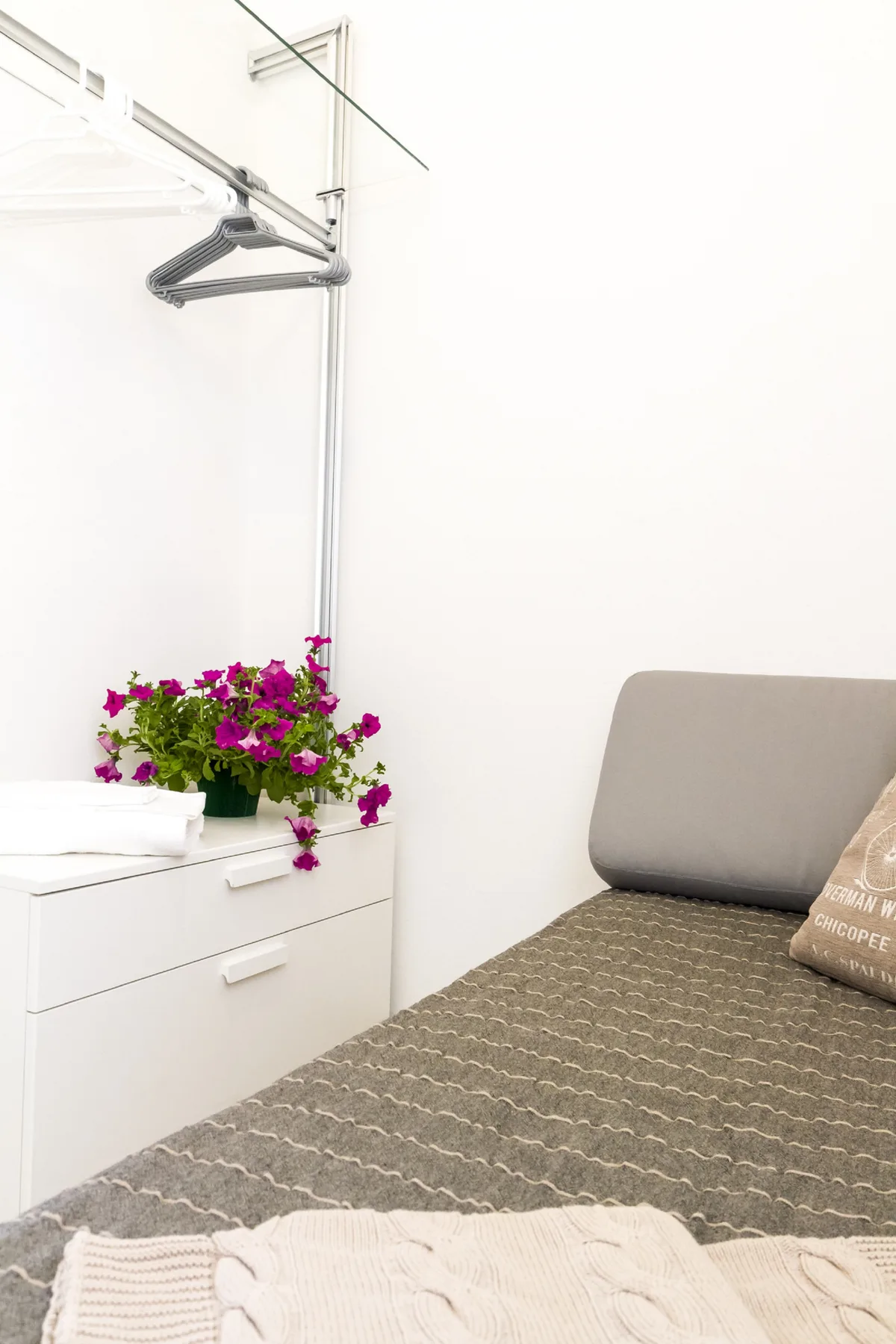 Un dormitorio con una cama gris, una cómoda blanca y una planta con flores.