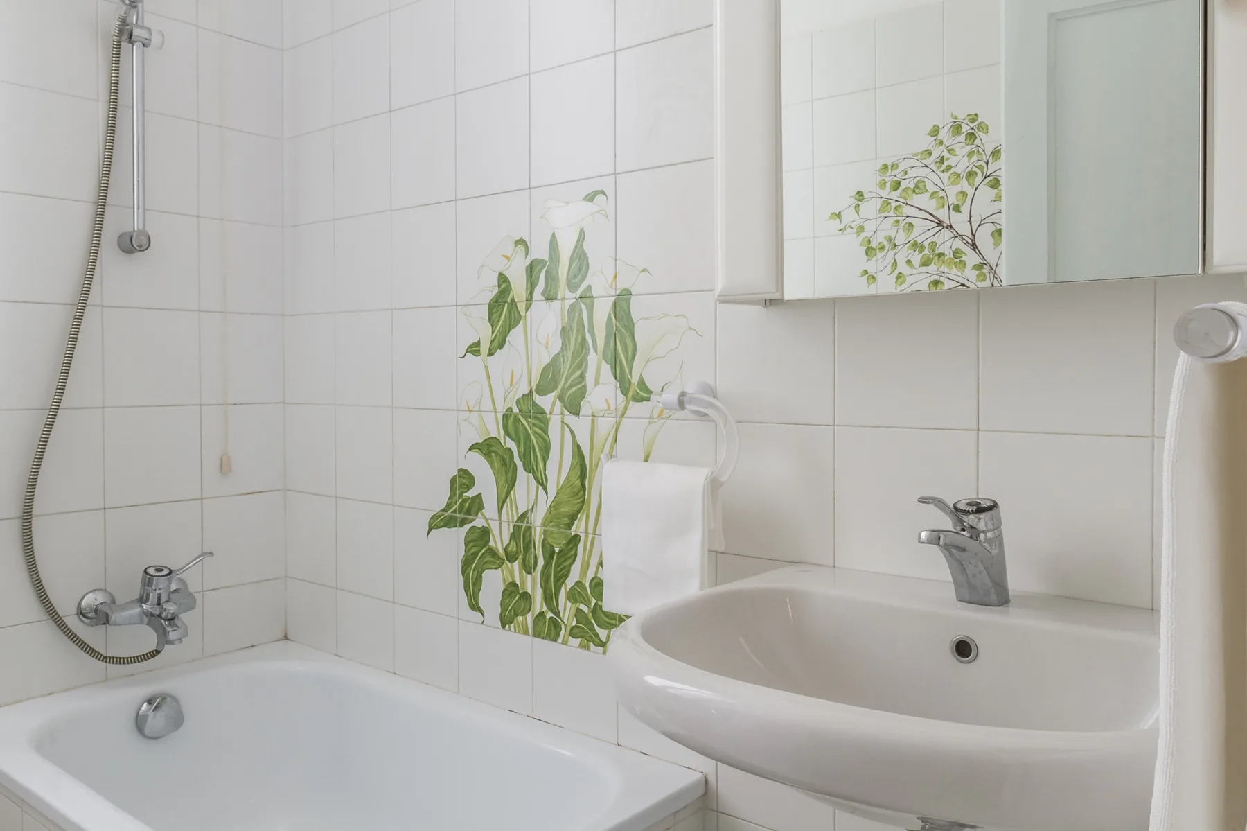 Un baño alicatado con bañera, lavabo y decoración vegetal en la pared.