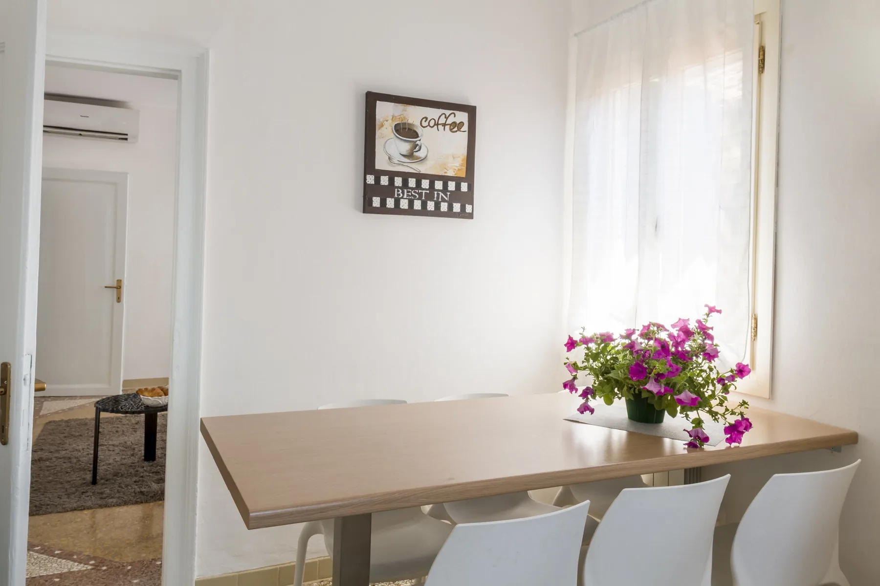 Comedor con una mesa rectangular, asientos blancos y flores junto a la ventana.