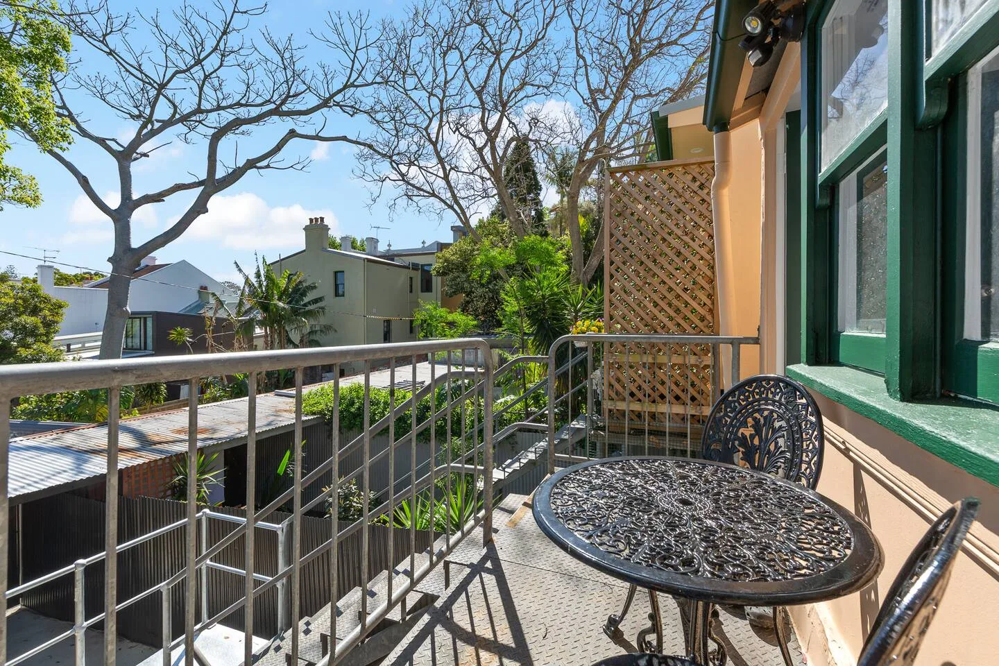 A small balcony with metal railing and outdoor seating overlooks leafy trees and nearby buildings.