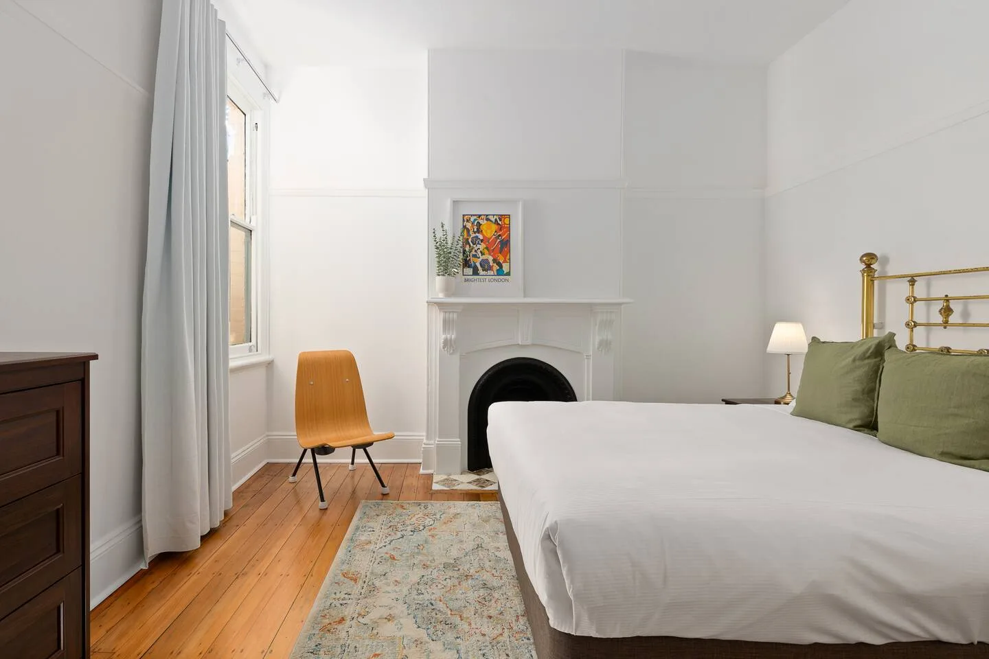 A bright bedroom with a bed, a fireplace, and a tall window.