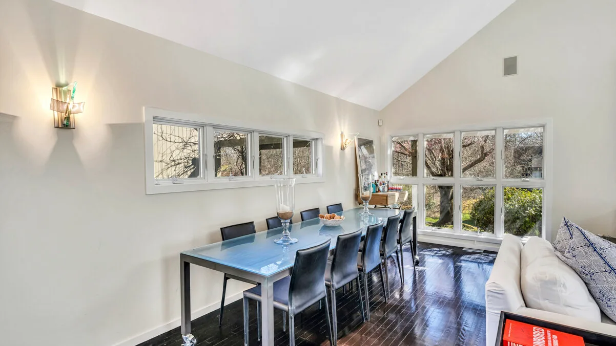 A bright dining area has a long table beside large windows and a seating area.