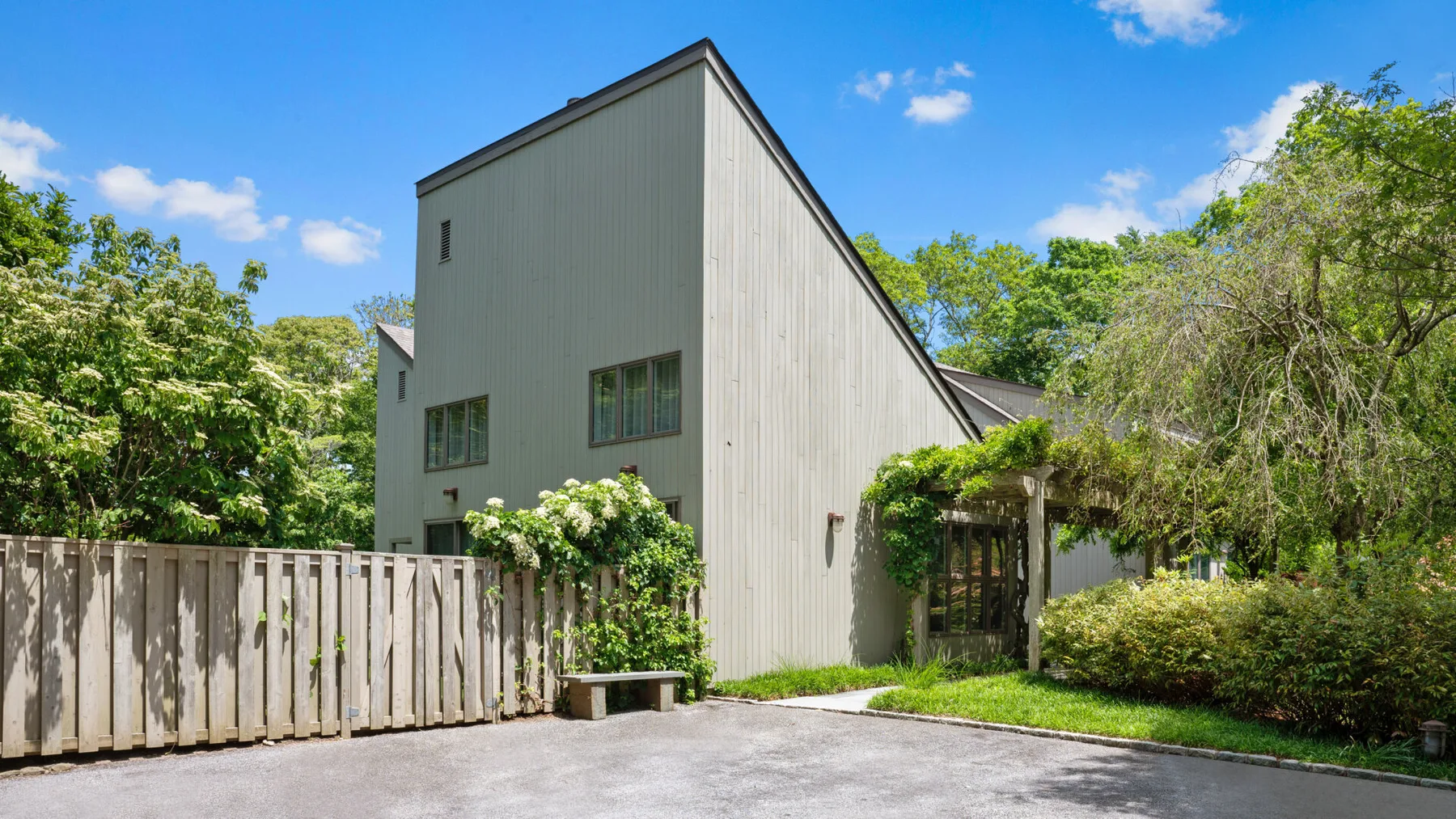 A tall-sided house stands behind a wooden fence, surrounded by greenery.