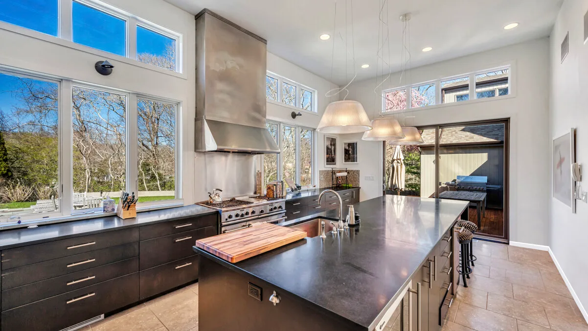 A bright kitchen with a long island and large windows overlooking trees.