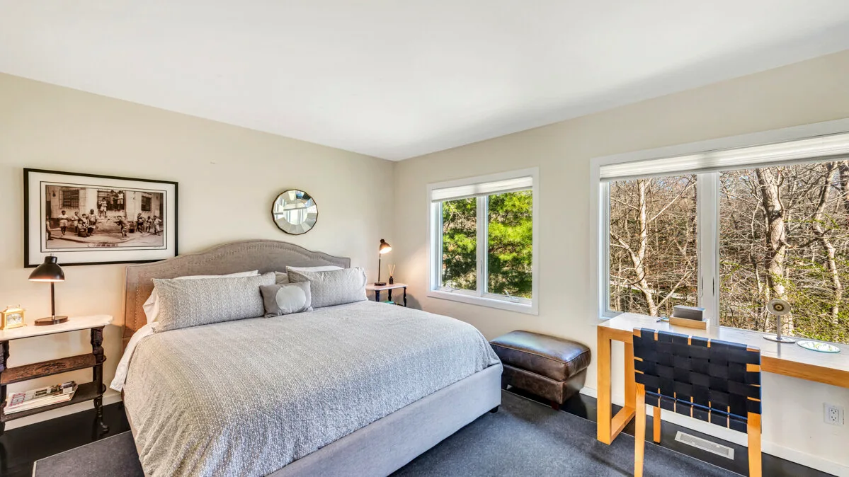A bedroom with a neatly made bed and desk beside large windows.