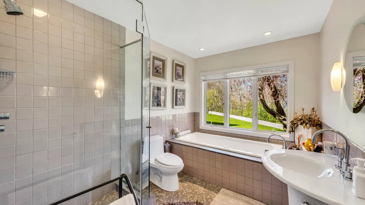 A bright bathroom with a glass shower, bathtub, and large window.