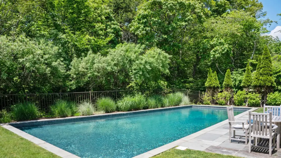 An outdoor swimming pool bordered by leafy trees and patio seating.