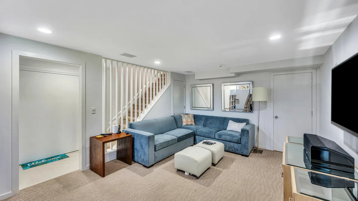 A living room with a blue sectional, white ottoman, and staircase.