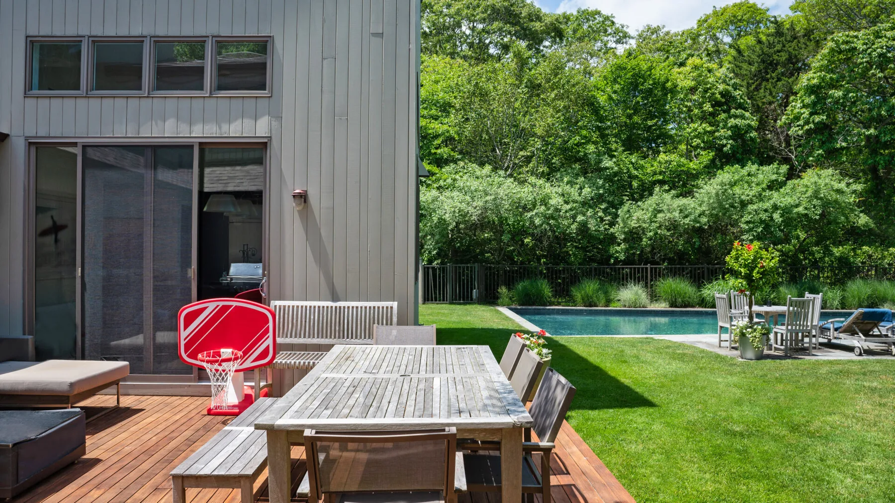 A wooden deck with an outdoor dining table overlooks a lawn and swimming pool.