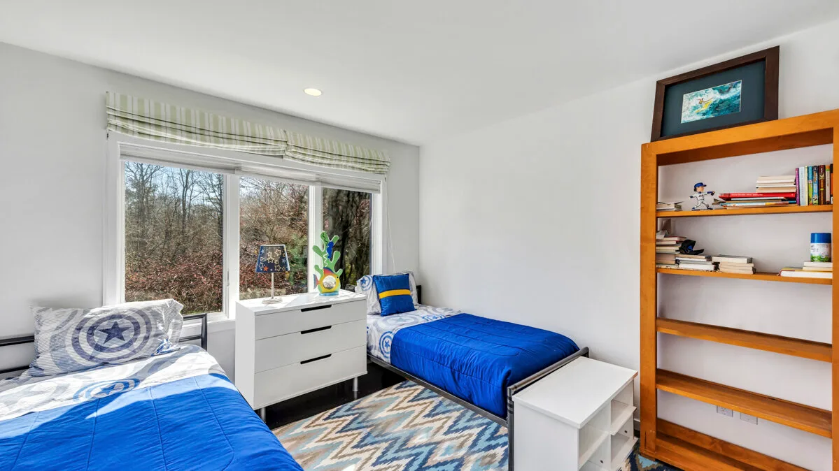 A bright bedroom with two beds, a window, and a wooden bookshelf.